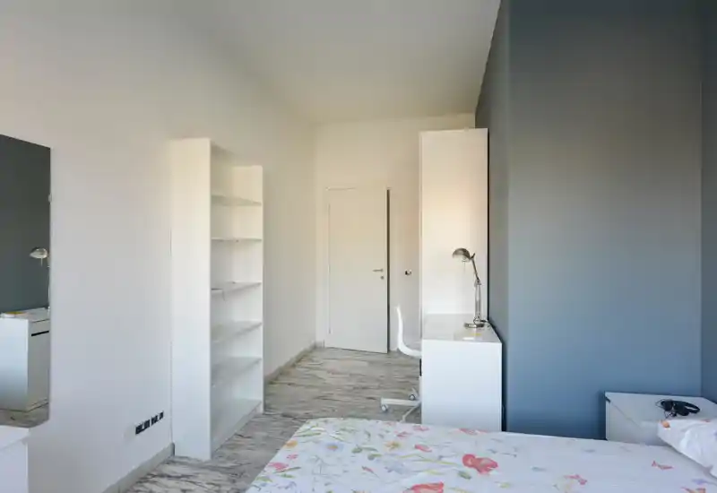 Dormitorio luminoso y minimalista con colcha floral, estantería blanca y un pequeño escritorio con lámpara. Paredes neutras y suelo efecto mármol que crean una atmósfera tranquila y aireada.