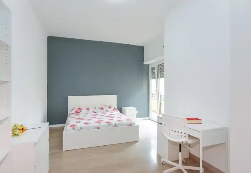Dormitorio luminoso y minimalista con cama blanca y ropa de cama floral, mesita de noche y escritorio junto a la ventana-balconera. Líneas limpias y luz natural crean un espacio aireado y acogedor, perfecto para anuncios.