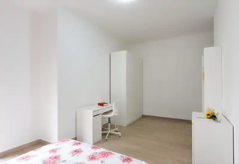 Dormitorio minimal y luminoso con mobiliario blanco: se aprecia la esquina de la cama con funda floral, un escritorio y un armario, espacio limpio y aireado ideal para alquiler pequeño o habitación de invitados.