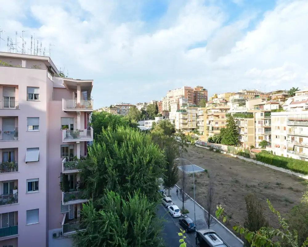 Vista luminosa desde un balcón hacia edificios vecinos, árboles y un terreno abierto; buena luz diurna y cielo despejado.