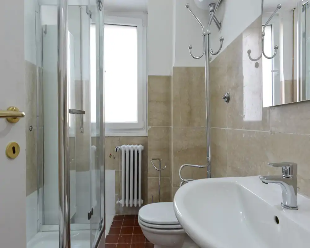 Baño compacto y limpio con lavabo, inodoro, mampara de ducha de vidrio y una ventana que aporta luz natural.