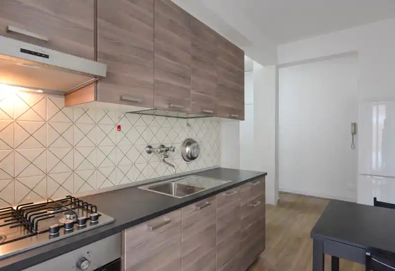 Cocina moderna equipada con muebles efecto madera, vitrocerámica a gas, fregadero de acero y salpicadero de azulejos geométricos. Espacio luminoso, ordenado y funcional, ideal para anuncios de apartamentos.