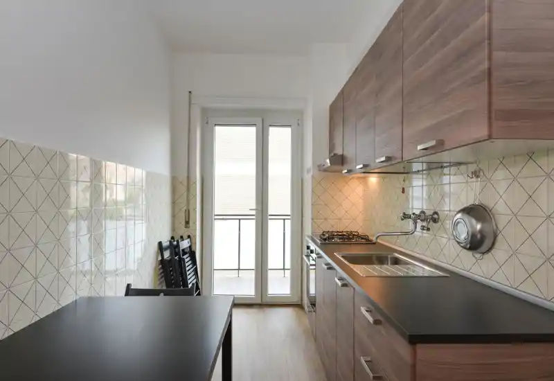 Cocina tipo pasillo moderna con muebles efecto madera, encimera negra, fregadero de acero inoxidable y placa de gas; la puerta al balcón aporta mucha luz natural.