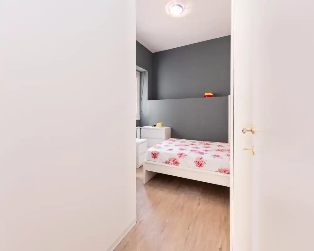 Dormitorio luminoso y ordenado con cama blanca y ropa de cama floral, pequeña mesita blanca y pared de acento gris; luz natural suave y decoración mínima crean un ambiente relajante.