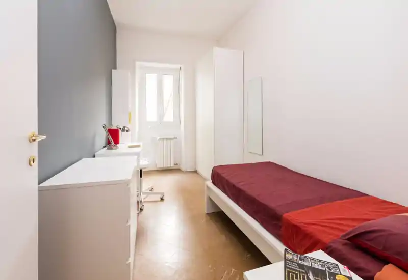 Camera da letto compatta e luminosa con letto singolo con biancheria rossa, scrivania vicino alla finestra e armadi bianchi dal design minimal. Linee pulite e luce naturale ideali per uno studente o occupante singolo.