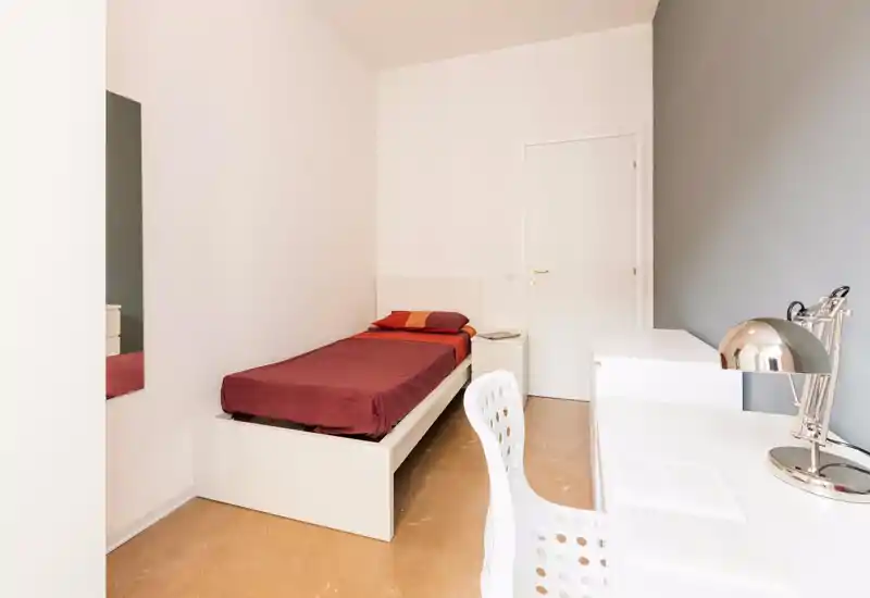 Camera singola minimal con letto rifatto, comodino e area scrivania luminosa. Pareti bianche e arredo semplice creano uno spazio tranquillo e ben illuminato ideale per studenti o affitti brevi.