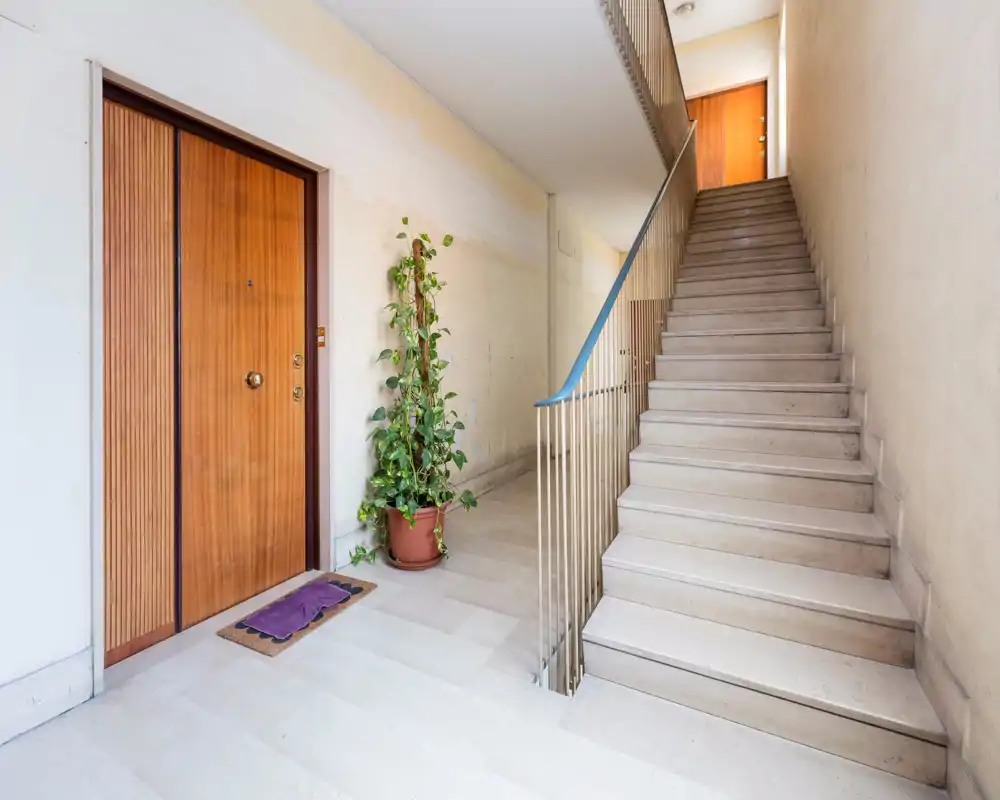Ingresso condominiale luminoso e pulito con porta d'ingresso in legno, vaso con pianta e scala che conduce ai piani superiori — ideale per descrivere il corridoio d'ingresso di un appartamento.