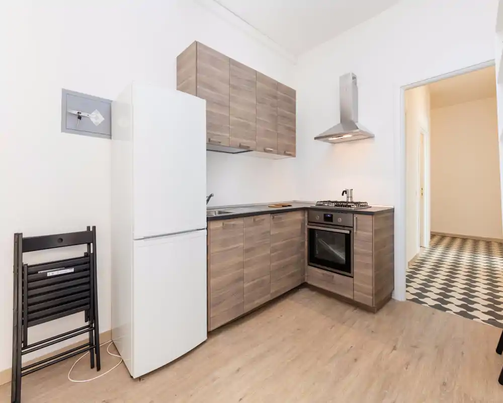 Cucina moderna compatta con pensili effetto legno, forno incassato, piano cottura a gas e frigorifero; tonalità neutre e pavimento in legno chiaro creano un ambiente caldo e funzionale.
