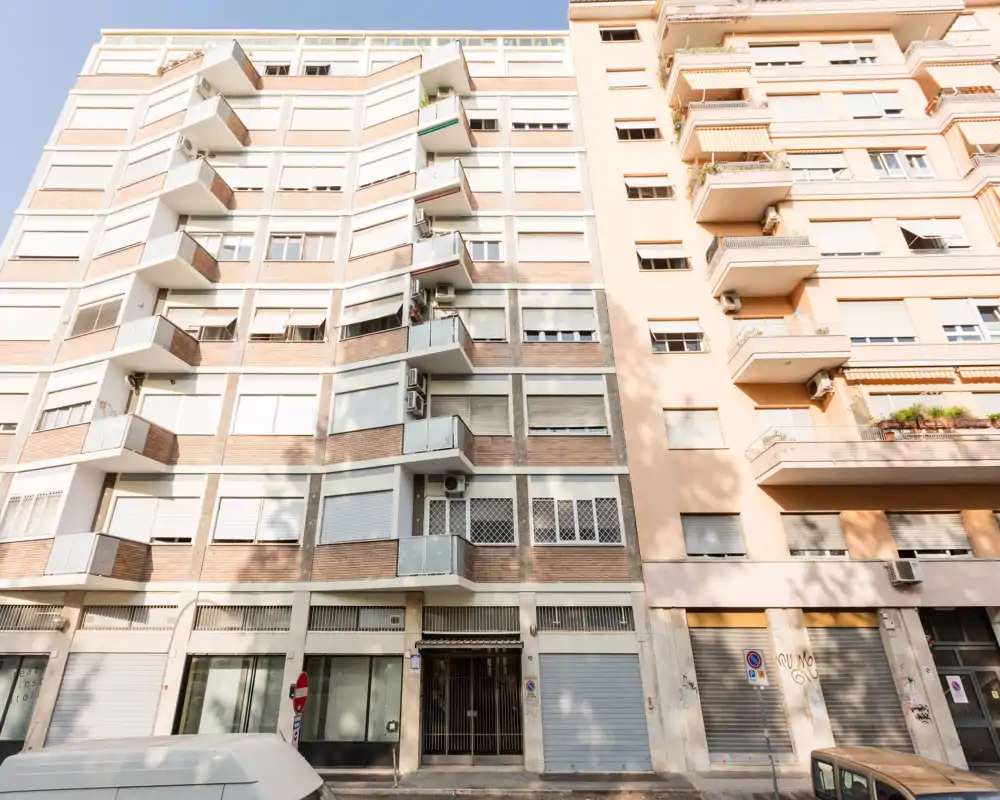 Vista della facciata esterna di un edificio residenziale di media altezza con balconi e negozi al piano terra; luce diurna intensa, adatta per mostrare l’ingresso o la posizione dell’appartamento.
