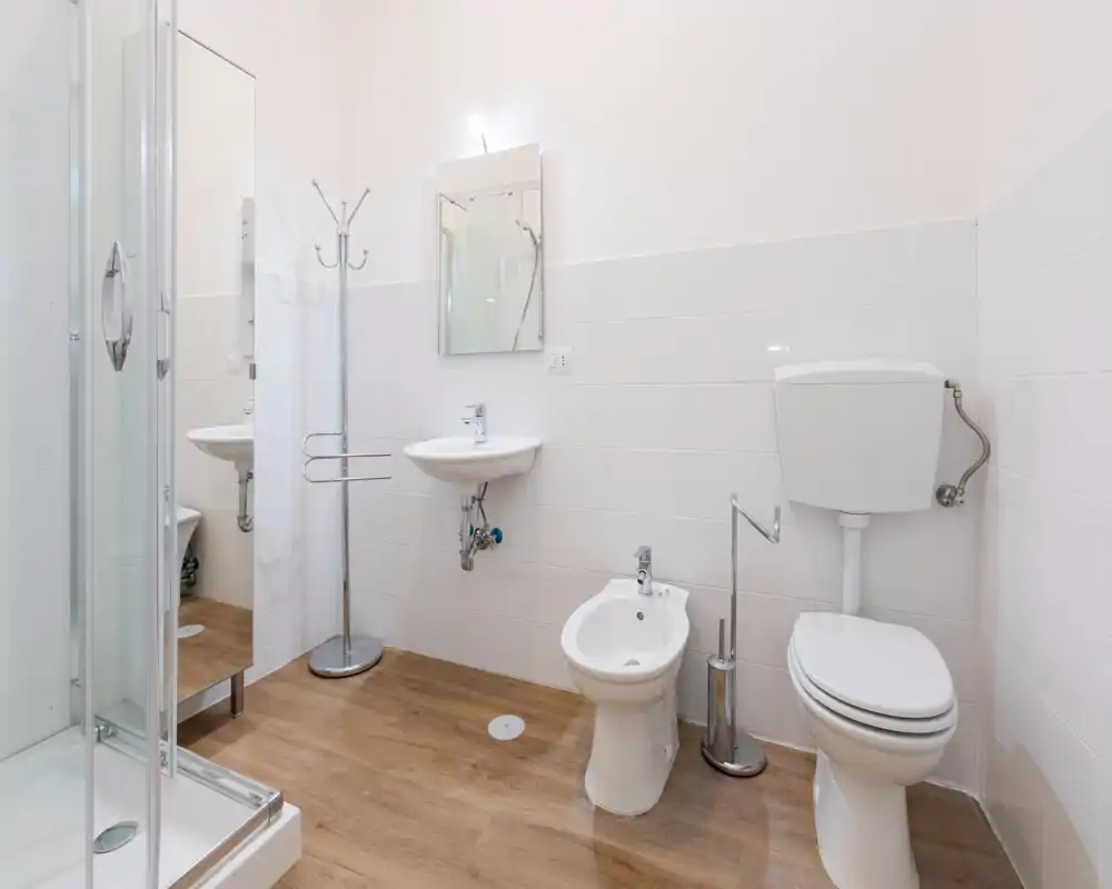 Bagno moderno e pulito con piastrelle bianche, box doccia, lavabo, bidet e WC su pavimento effetto legno; ambiente luminoso e minimal.