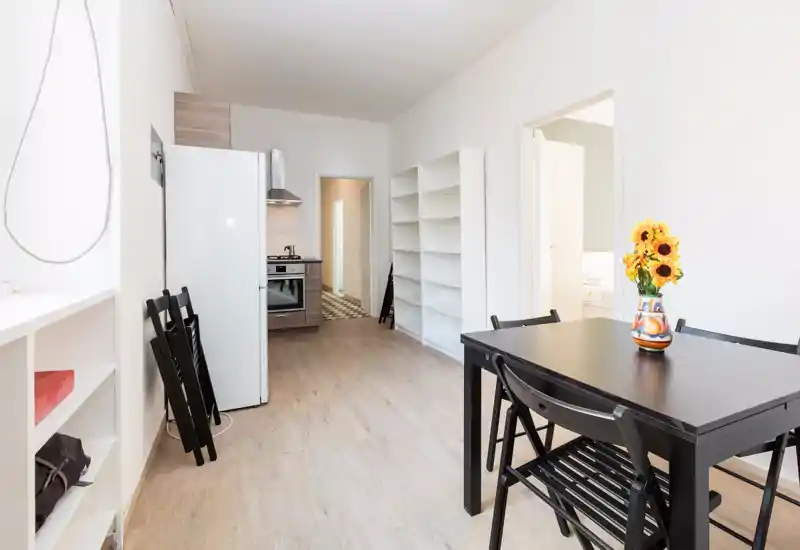Cucina compatta e luminosa con piccolo tavolo da pranzo e sedie, frigorifero e forno; stile pulito e minimale con girasoli decorativi sul tavolo.