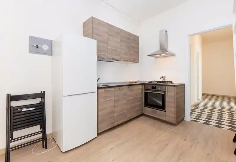 Cucina moderna compatta con pensili effetto legno, forno incassato, piano cottura a gas e frigorifero; tonalità neutre e pavimento in legno chiaro creano un ambiente caldo e funzionale.