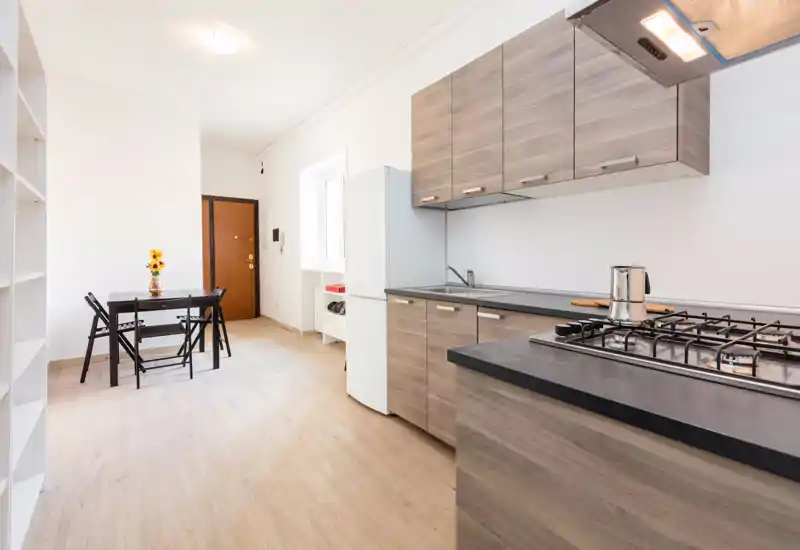 Cucina moderna e luminosa con pensili effetto legno, piano cottura a gas, frigorifero e piccolo tavolo da pranzo — perfetta per un appartamento compatto.