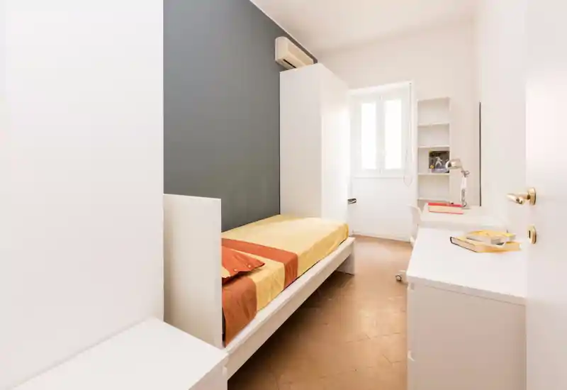 Compact, neat bedroom with a single bed, minimal white furniture and a bright window that floods the room with natural light.