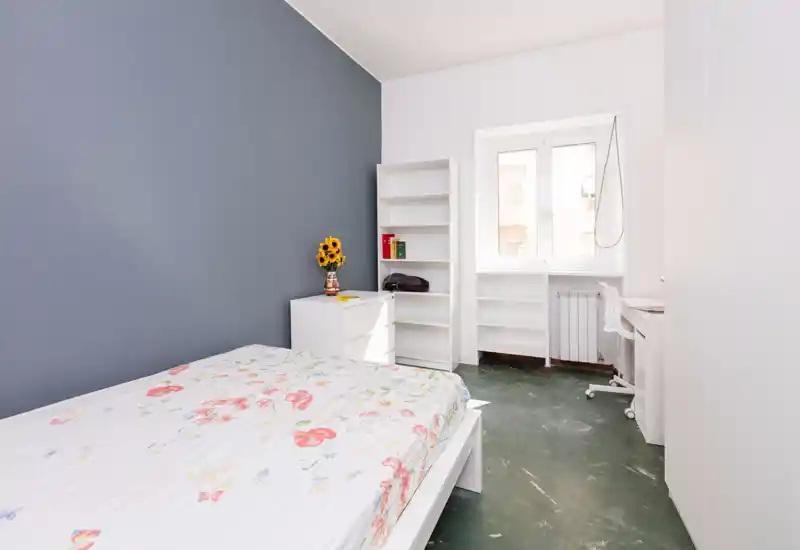 Camera da letto luminosa e minimale con letto a fantasia floreale, mobili bianchi e parete grigia accentata. La luce naturale dalla finestra e il vaso di girasoli creano un’atmosfera fresca e accogliente.