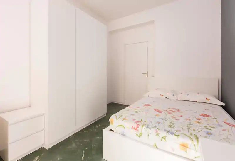 Camera da letto minimal e ben illuminata con un armadio bianco e letto con copertura floreale. Linee pulite e toni neutri donano un’atmosfera rilassante.