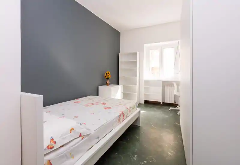 Camera da letto luminosa e minimale con un letto bianco in primo piano, copriletto floreale, scaffali e una finestra che porta luce naturale. La parete grigia d'accento e l'arredamento semplice creano un aspetto pulito e moderno.