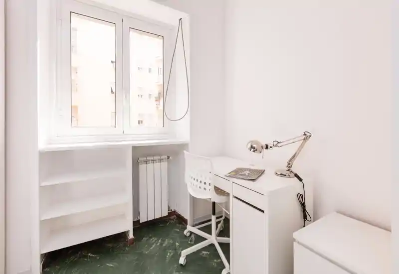 Stanza luminosa e minimal con una piccola scrivania bianca e sedia accanto a una grande finestra; ideale come angolo studio compatto con luce naturale e una lampada da tavolo elegante.
