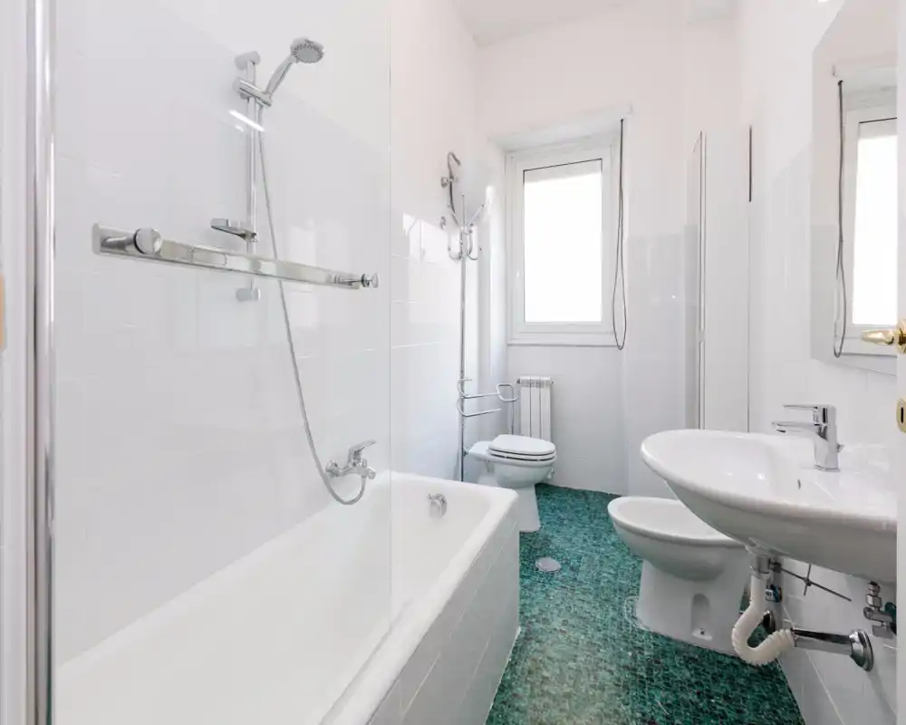 Bright, modern bathroom with bathtub, shower head, toilet and sink. Clean white tiles and a small window provide natural light.