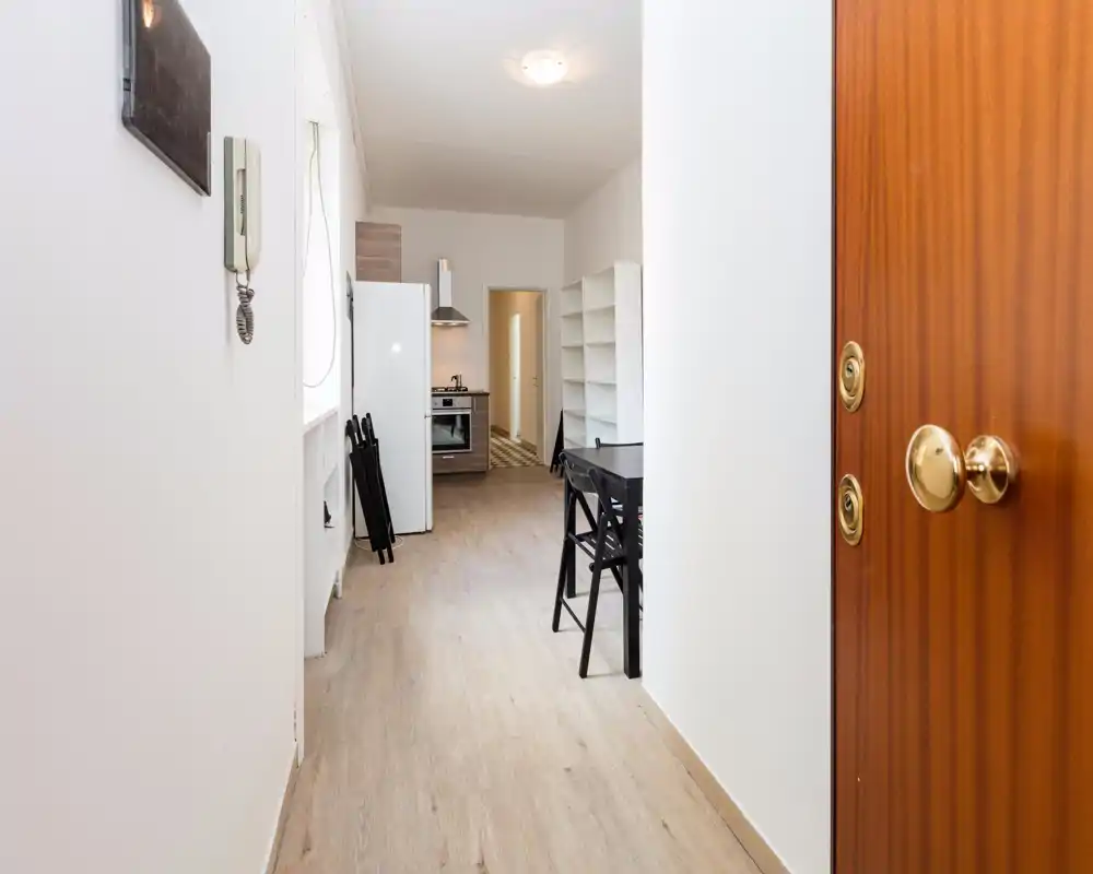 Bright entry hallway with a view into a compact kitchen; light wood flooring, white walls and a small dining table create a clean, welcoming arrival space.