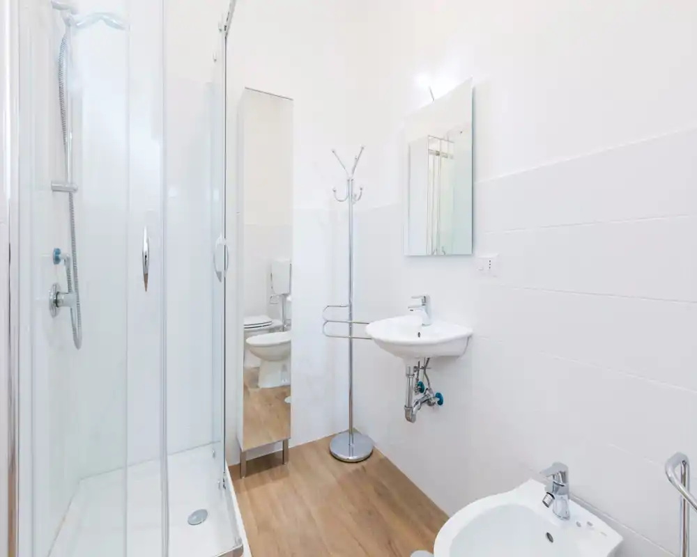 Bright, modern bathroom with a glass shower enclosure, wall-mounted sink and mirror; light tiles and wood-effect floor create a clean, minimalist look.