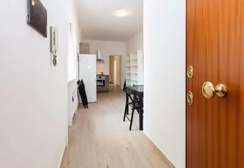 Bright entry hallway with a view into a compact kitchen; light wood flooring, white walls and a small dining table create a clean, welcoming arrival space.