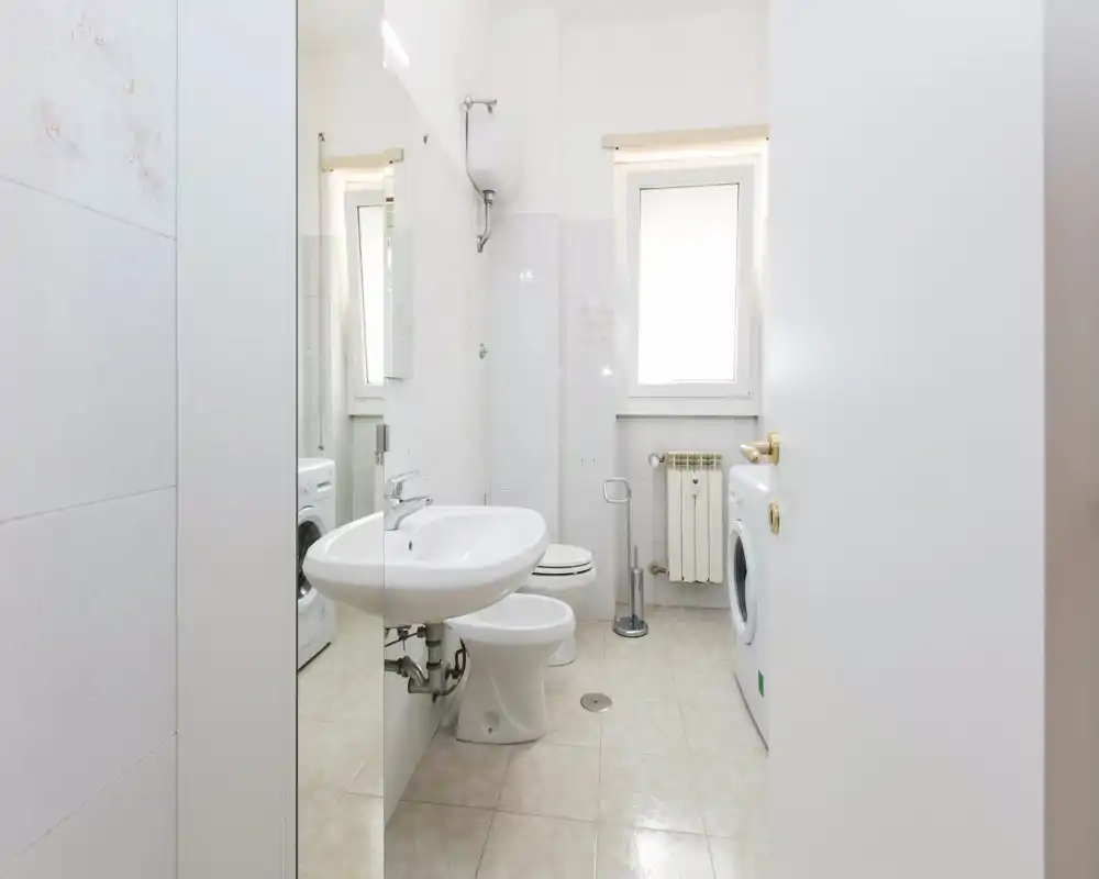 Bagno compatto e luminoso con piastrelle bianche, lavabo, wc e lavatrice; pulito e ben illuminato, ideale per un appartamento piccolo.