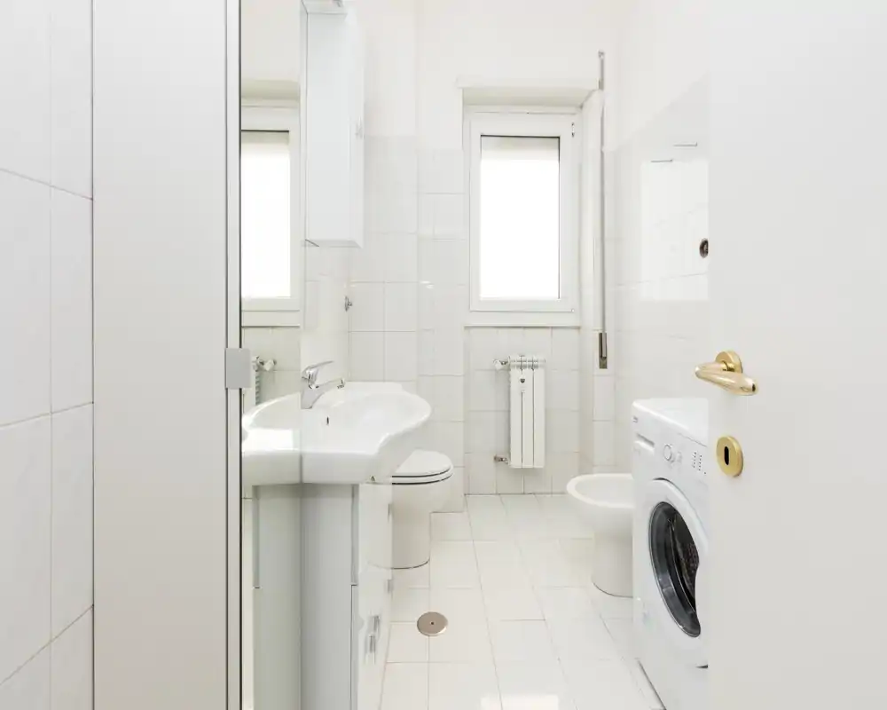 Bagno compatto e luminoso con lavabo, WC, bidet e lavatrice; piastrelle chiare e finestra che garantisce luce naturale.