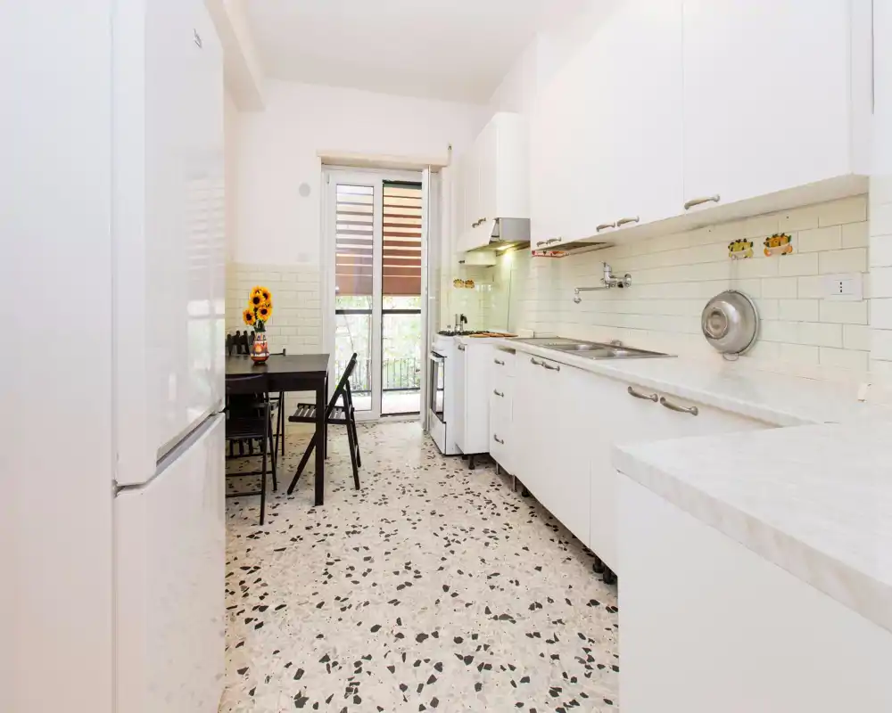 Cucina compatta e luminosa con pensili bianchi, piani effetto marmo e un piccolo tavolo da pranzo vicino alla porta del balcone — moderna e ben illuminata.