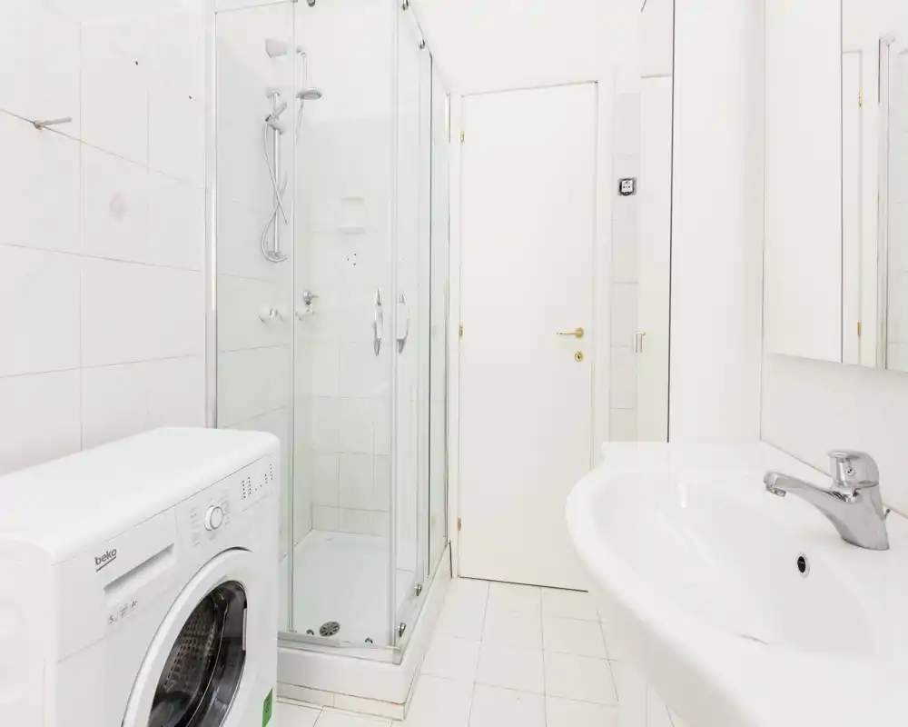 Bagno luminoso e pulito con box doccia in vetro, lavabo e lavatrice. Lo spazio è prevalentemente bianco con piastrelle e soffione doccia cromato moderno.