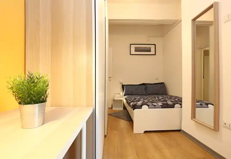 Bright, neatly staged bedroom with a double bed, bedside table and decorative textiles; warm wooden floor and a full-length mirror create a welcoming, functional space.