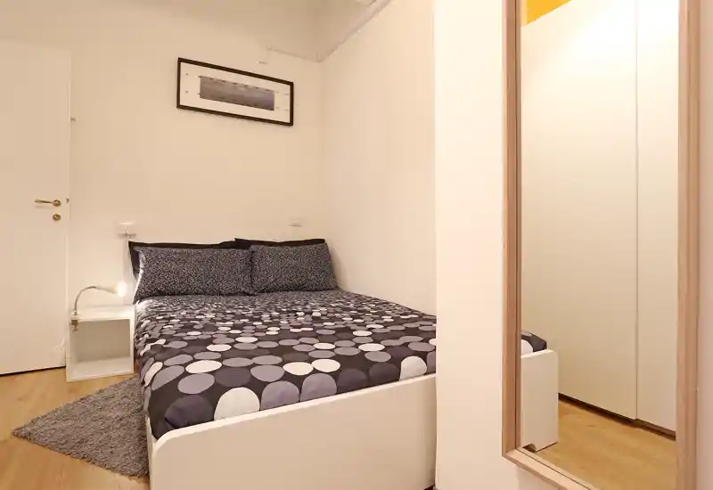 Bright, compact bedroom with a double bed, patterned bedding, bedside table and wall art. Clean, modern styling and a full-length mirror make the room feel larger and welcoming.