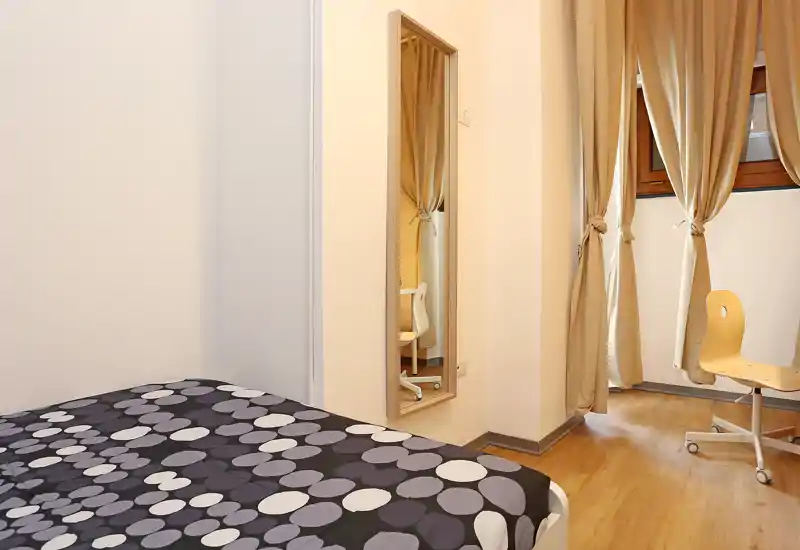 Tidy bedroom corner with a patterned double bed, warm wooden floors and full-length mirror; soft beige curtains let in warm light creating a cozy atmosphere.