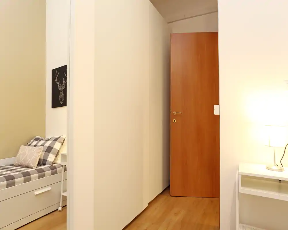 Rincón de dormitorio compacto y ordenado con una cama individual con ropa a cuadros, cojines decorativos y un cajón integrado. Iluminación cálida y paredes claras que aportan una sensación acogedora, perfecto para un apartamento pequeño o habitación de invitados.