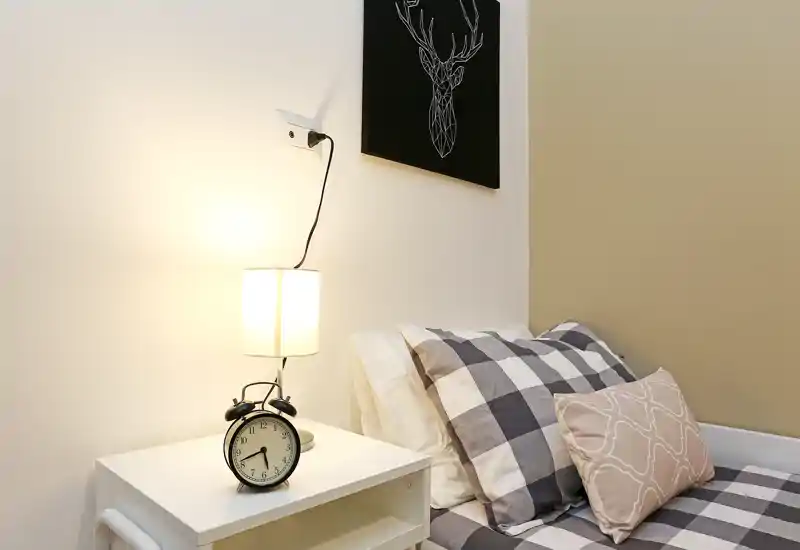 Rincón de dormitorio individual acogedor y bien iluminado con mesita, lámpara y cojines estampados. Tonos neutros y arreglo ordenado ideales para anuncios de alquiler.