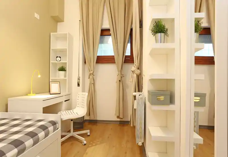 Dormitorio compacto y bien decorado con cama individual, escritorio y estanterías; iluminación cálida y cortinas neutras que crean un espacio acogedor y funcional.
