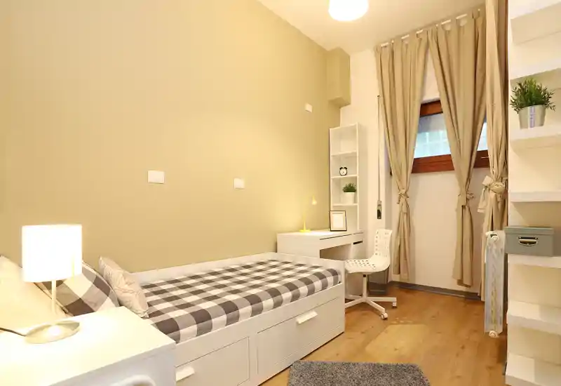 Dormitorio individual luminoso y ordenado con cama a cuadros, lámpara de noche, cajones integrados y pequeño escritorio junto a la ventana — ideal para estudiante o persona sola.