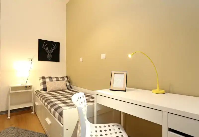 Dormitorio compacto y bien iluminado con cama individual de funda a cuadros, lámpara, escritorio y silla — ideal para estudiantes o estancias cortas.