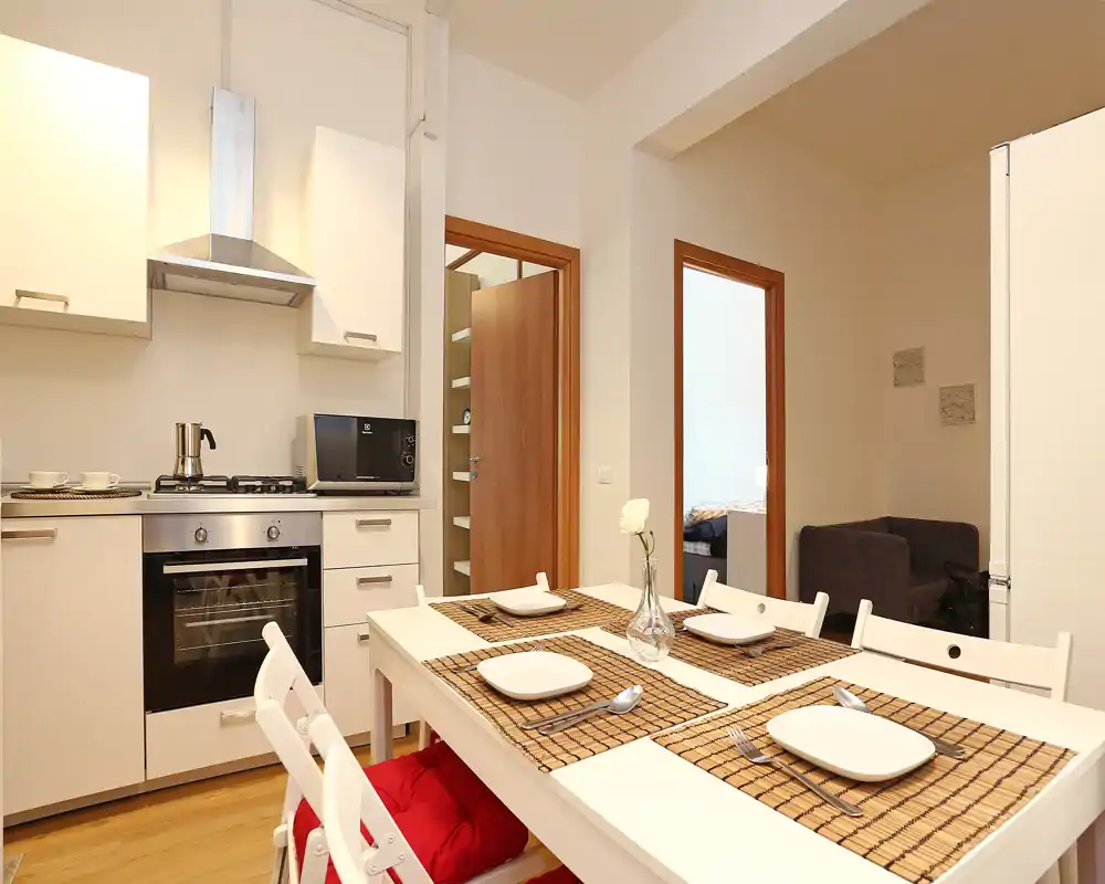 Cocina moderna abierta y luminosa con mesa puesta para cuatro, mobiliario blanco, horno y microondas empotrados — un espacio acogedor y bien iluminado, perfecto para comidas informales.