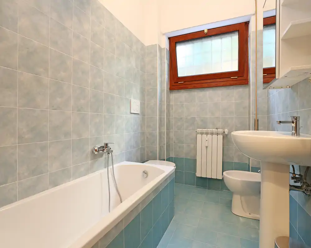 Baño limpio y luminoso con bañera, lavabo de pedestal e inodoro; azulejos azul claro y ventana esmerilada que aportan privacidad y sensación de frescura.