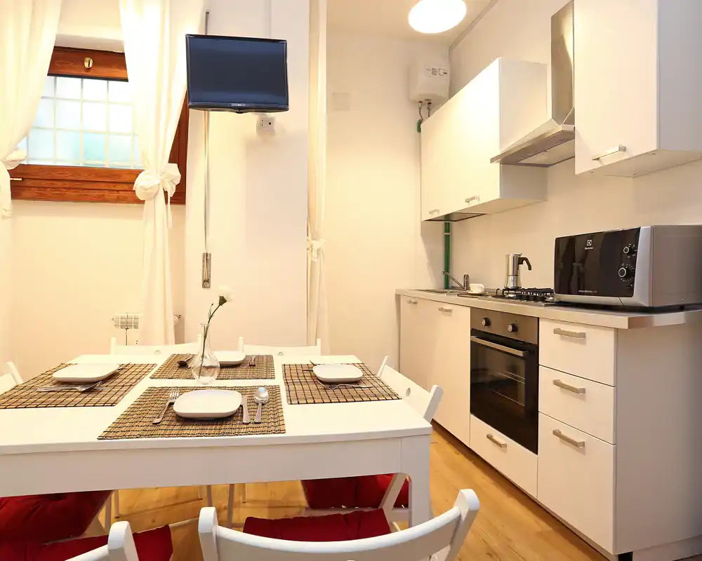 Cocina moderna y compacta con armarios blancos, horno empotrado, microondas y mesa para cuatro — luminosa y acogedora, ideal para grupos pequeños.