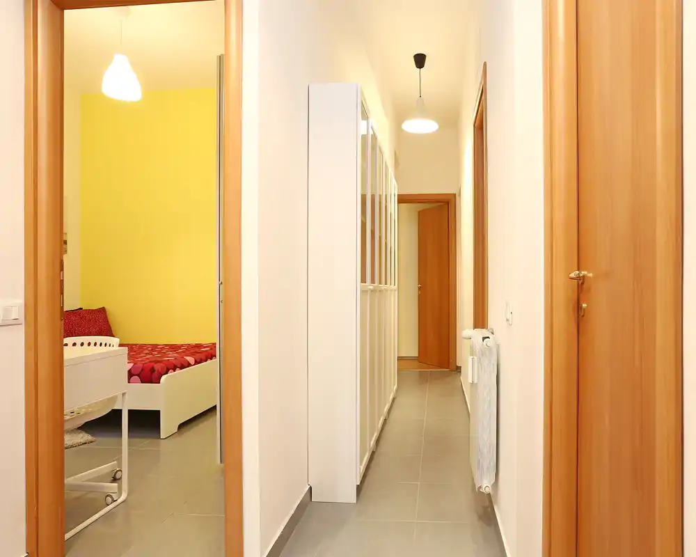 Pasillo luminoso con puertas de madera y radiador; se intuye una habitación con pared amarilla a través de la puerta abierta.