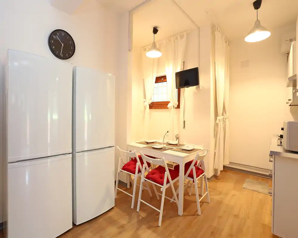 Cocina-comedor compacta y luminosa con mesa blanca, cuatro sillas plegables con cojines rojos, dos frigoríficos y lámparas colgantes cálidas — perfecta para un apartamento acogedor.