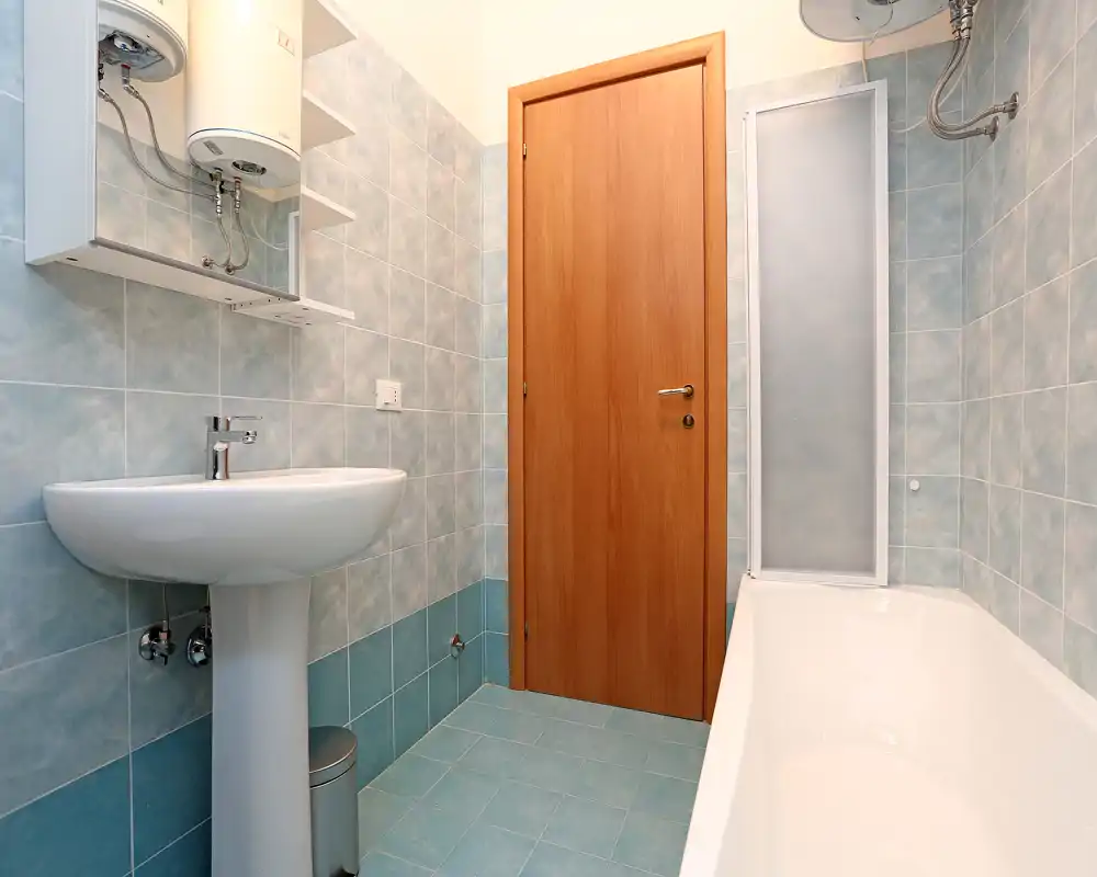 Baño compacto y limpio con bañera, lavabo de pedestal y azulejos azulados; luminoso y bien cuidado.