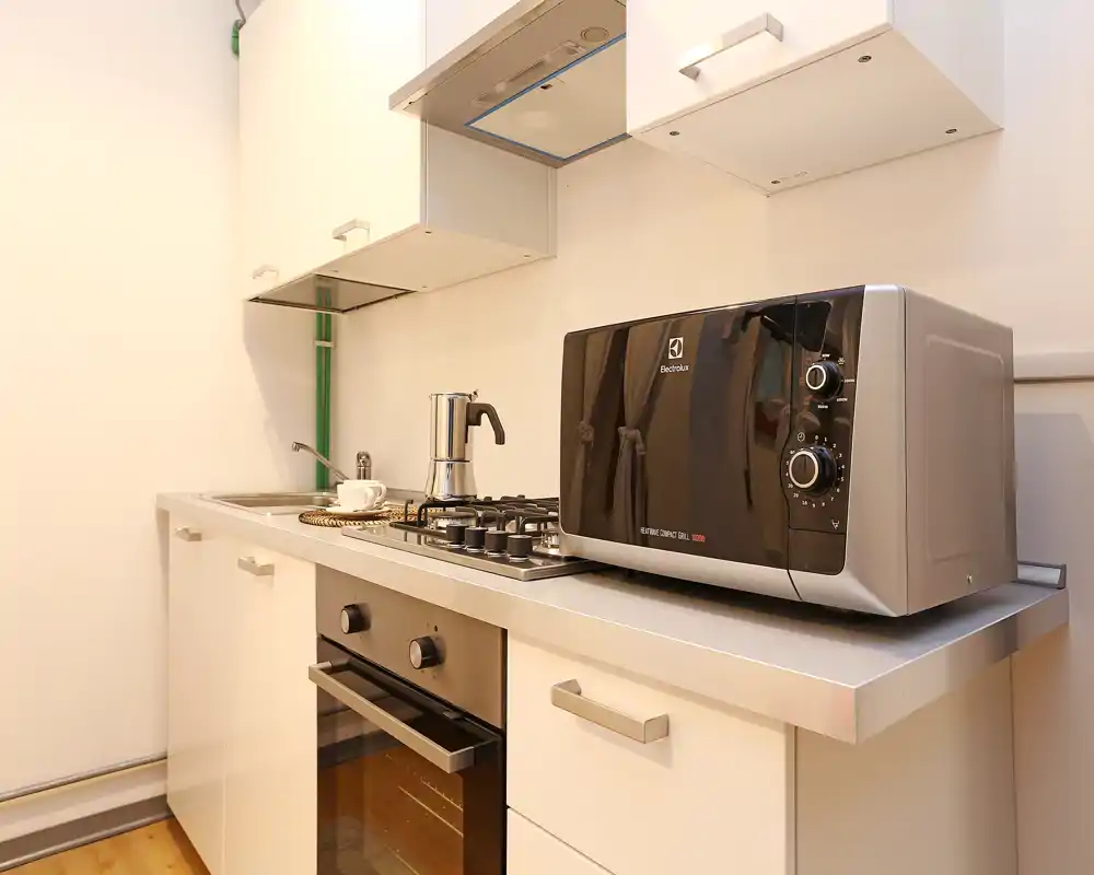 Cocina moderna y compacta con electrodomésticos inox, horno empotrado, placa de gas y microondas en la encimera; superficies limpias y luminosas, ideal para alquileres temporales.