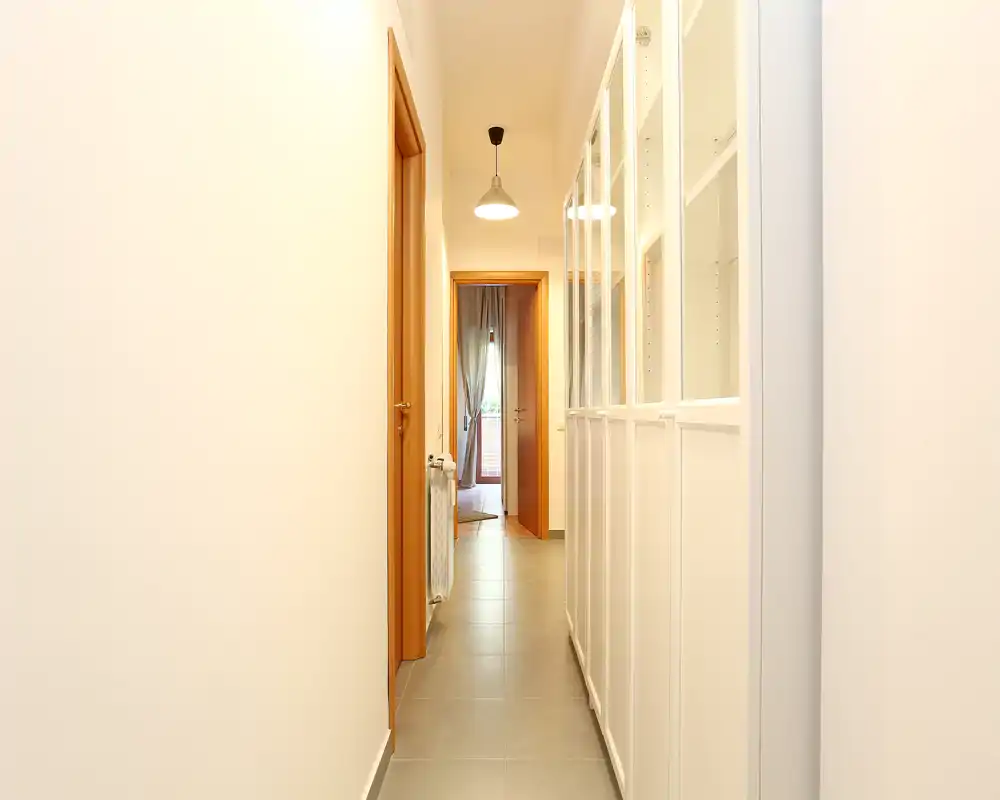 Pasillo de entrada luminoso y estrecho con suelo de baldosas, armario empotrado a la derecha y puertas de madera hacia las habitaciones interiores — bien iluminado y ordenado.
