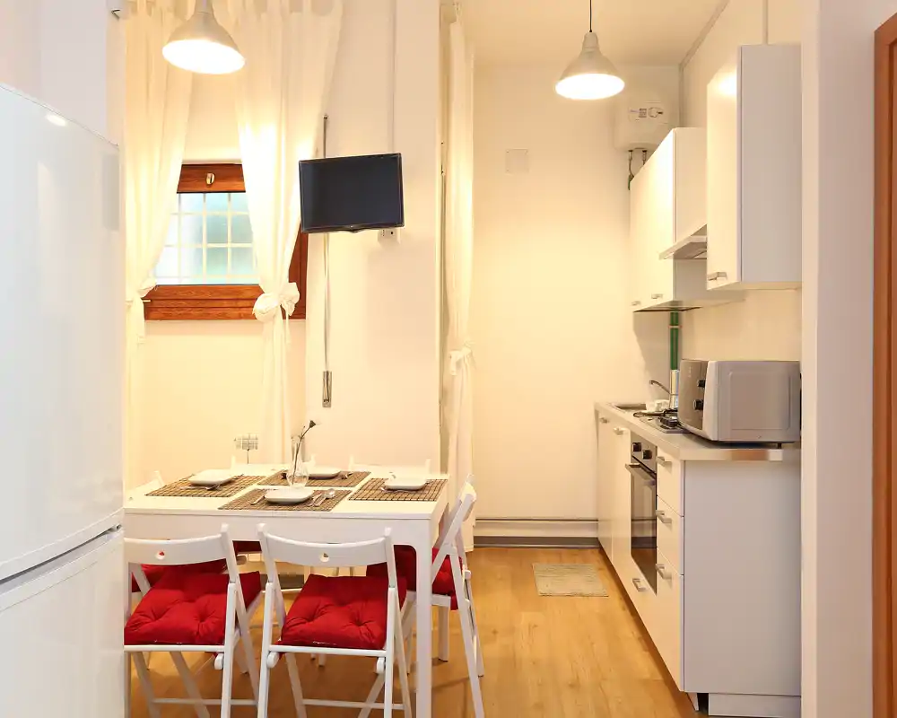 Cocina moderna y compacta con mesa de comedor para cuatro; muebles blancos, suelo de madera y luz cálida que crean un ambiente acogedor.