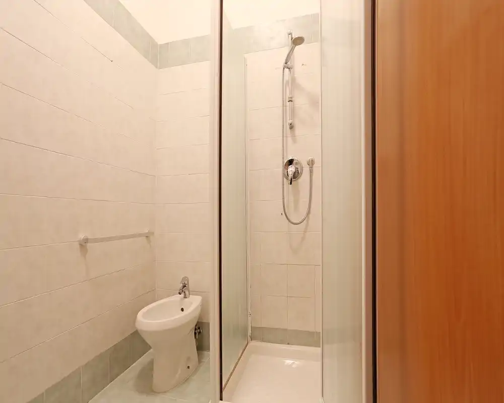 Baño alicatado y compacto con ducha en esquina y bidé; azulejos claros y mampara de vidrio que ofrecen un espacio limpio y funcional.