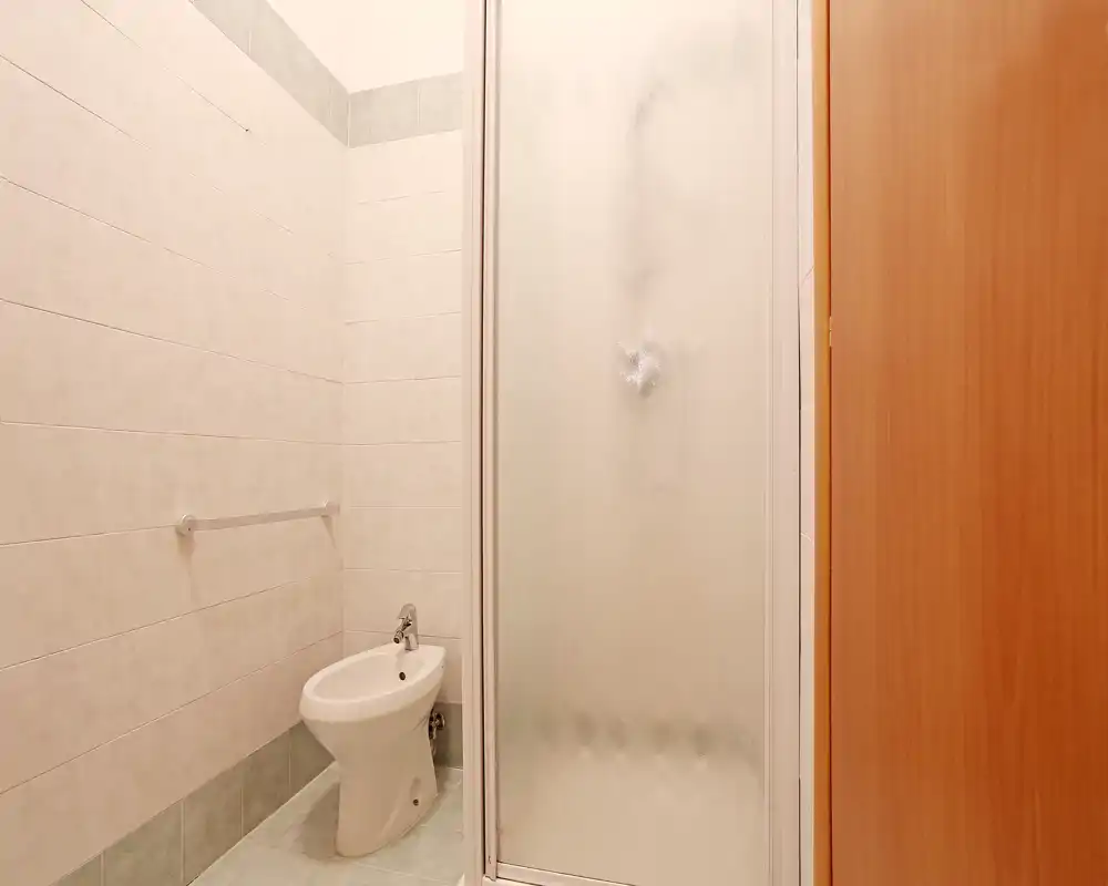 Baño pequeño y limpio con bidé y ducha cerrada de vidrio esmerilado; paredes alicatadas en tonos neutros y puerta de madera visibles.