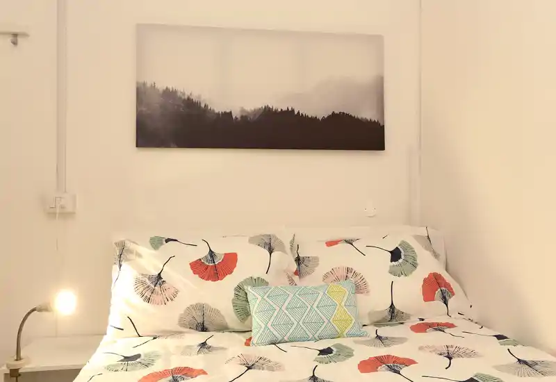 Camera da letto luminosa e accogliente con biancheria a fantasia, cuscino decorativo e lampada da comodino; pareti neutre e quadro paesaggistico sopra il letto donano tranquillità.